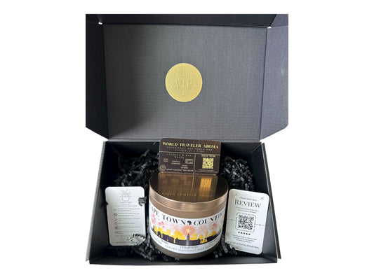 A black gift box with a gold VIP sticker on the lid inside the box a gold candle tin labeled "Cape Town Countdown" with a candle care card instructions, a please review us card, and World Traveler Aroma business card on a white background.