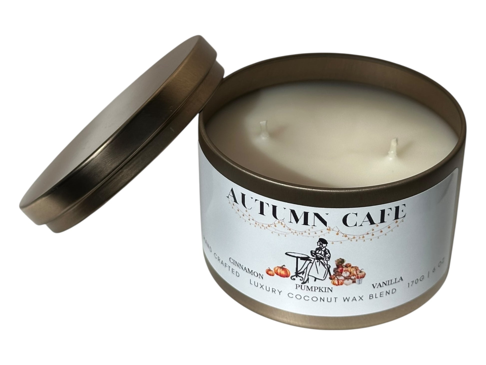 A gold candle tin labeled 'Autumn Cafe' with a decorative label lid removed showing two candle wicks and a smooth white candle top on a white background.
