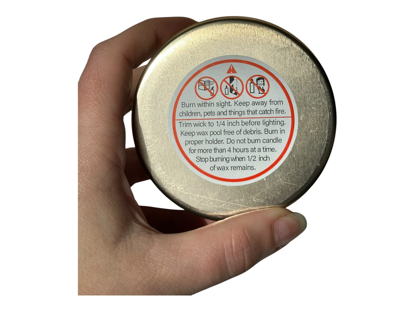 A hand holding a gold candle tin with a warning label on a white background.
