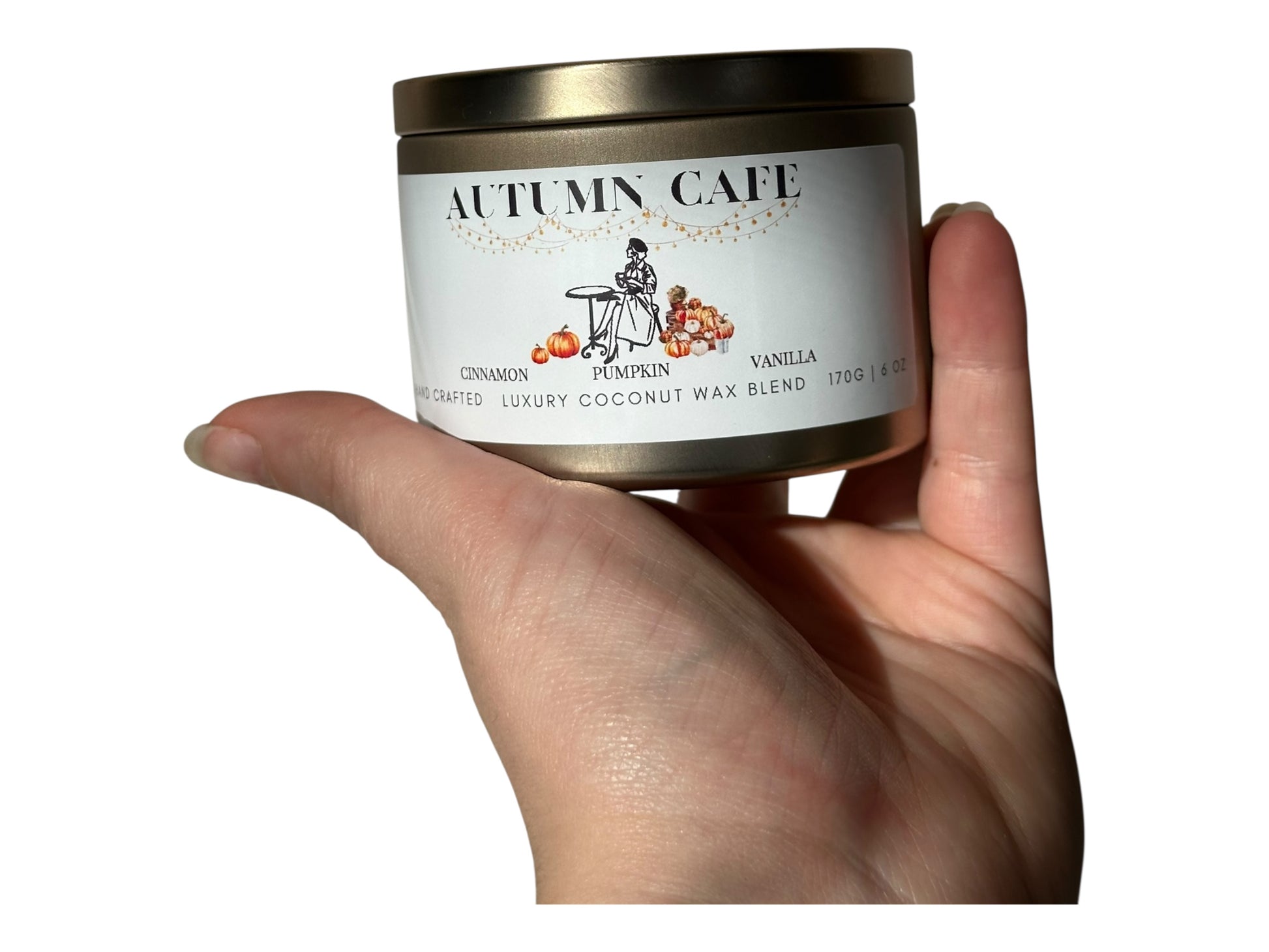 A hand holding a gold candle tin labeled 'Autumn Cafe' with a white background.