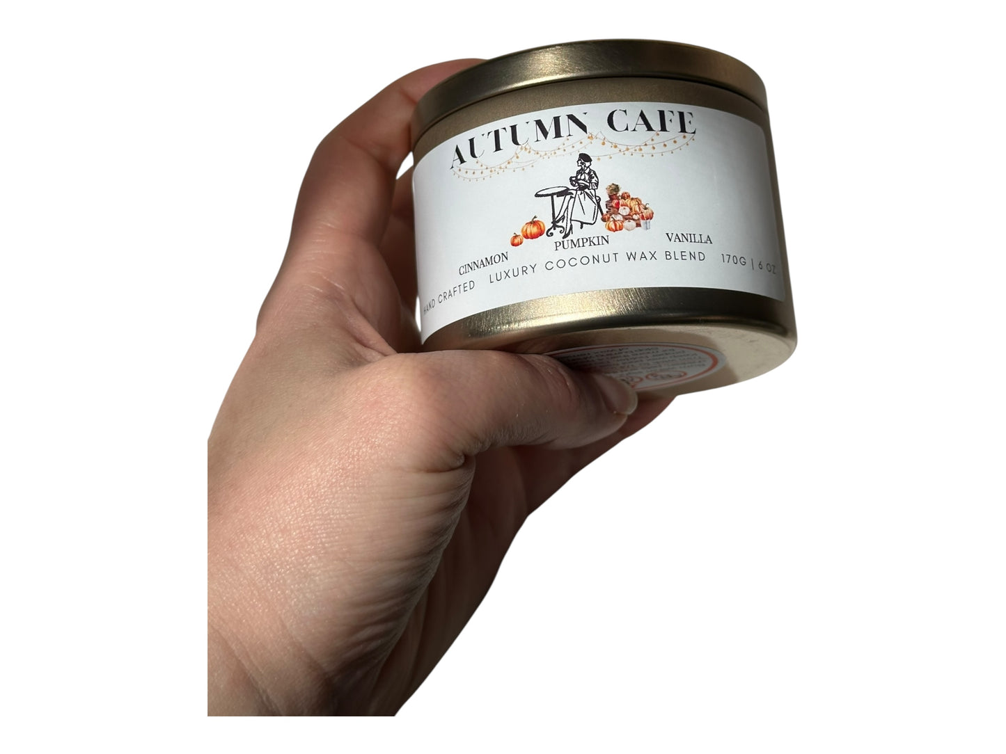 A hand holding a gold candle tin labeled 'Autumn Cafe' against a white background.