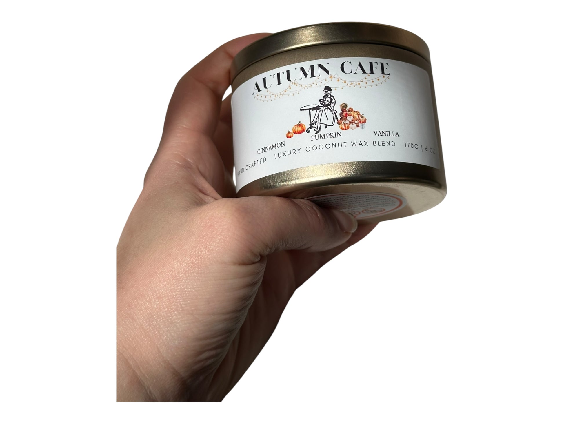 A hand holding a gold candle tin labeled 'Autumn Cafe' against a white background.