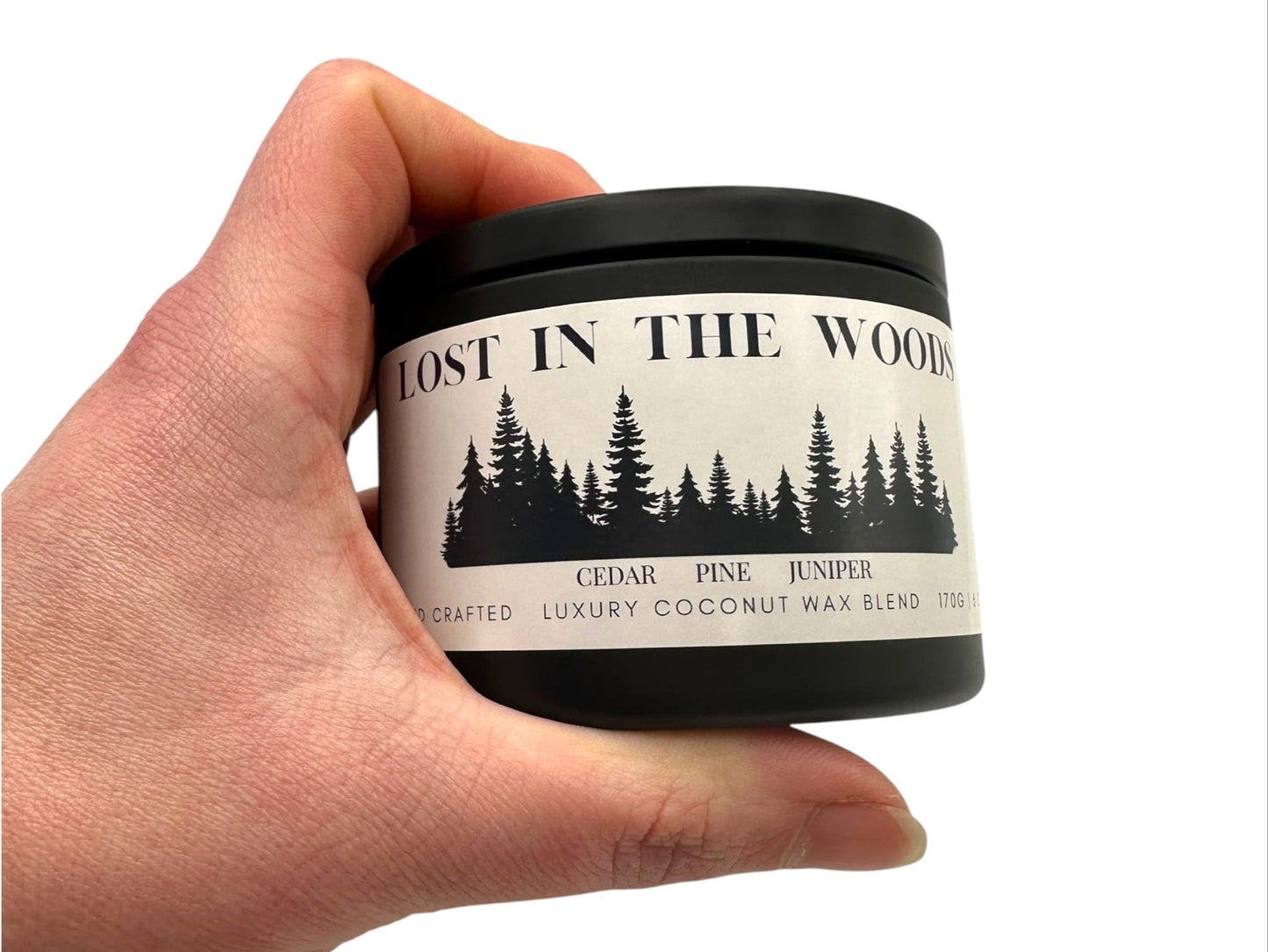A hand holding a black candle tin labeled "Lost in the Woods" on a white background.