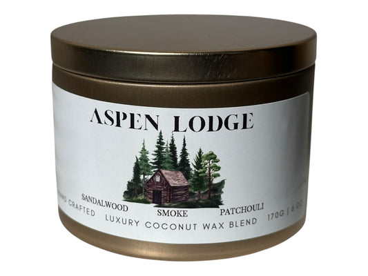 A gold candle tin labeled 'Aspen Lodge' with a cabin and tree design on the label on a white background.