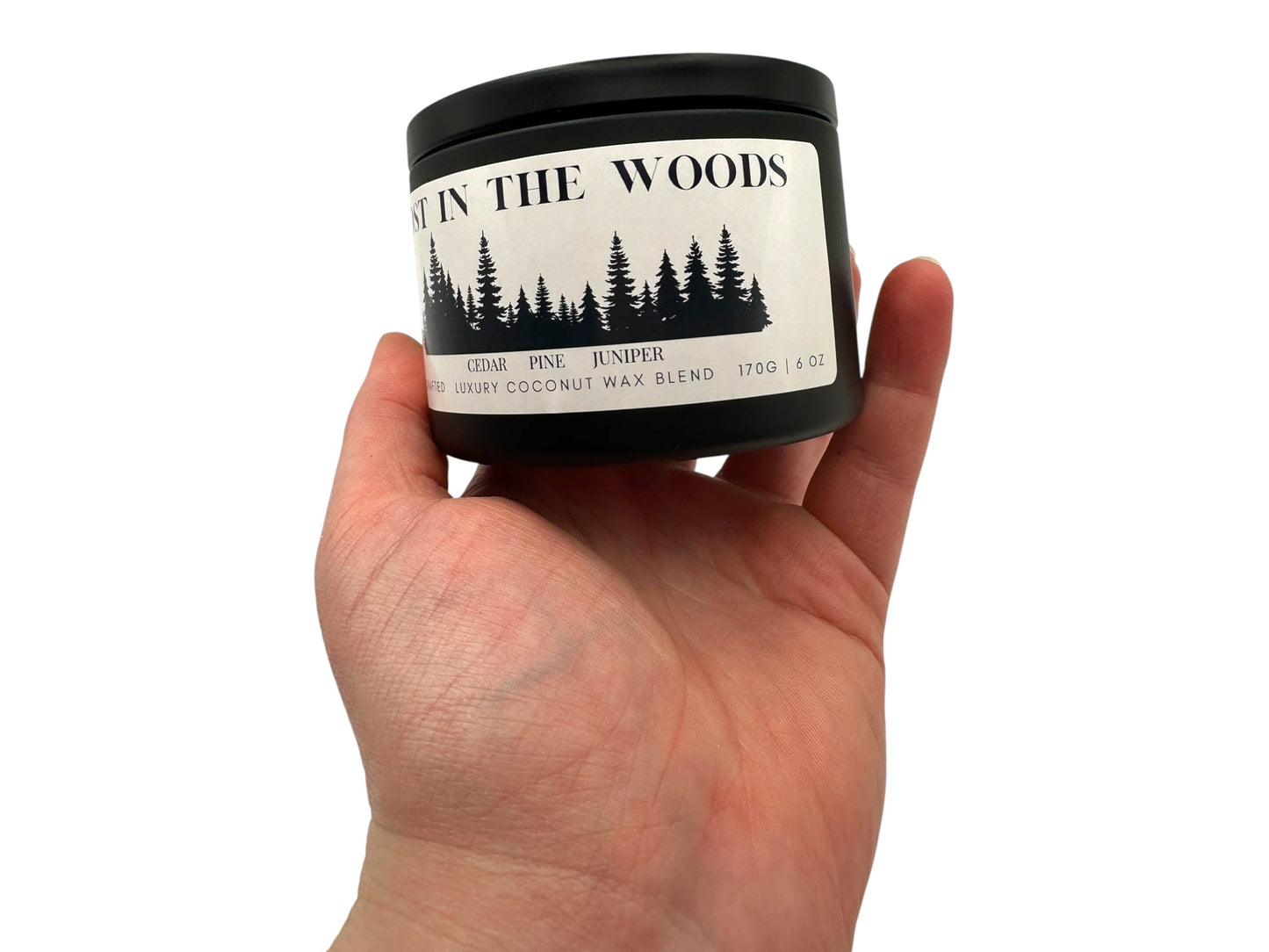A hand holding a black candle tin labeled "Lost in the Woods" on a white background.