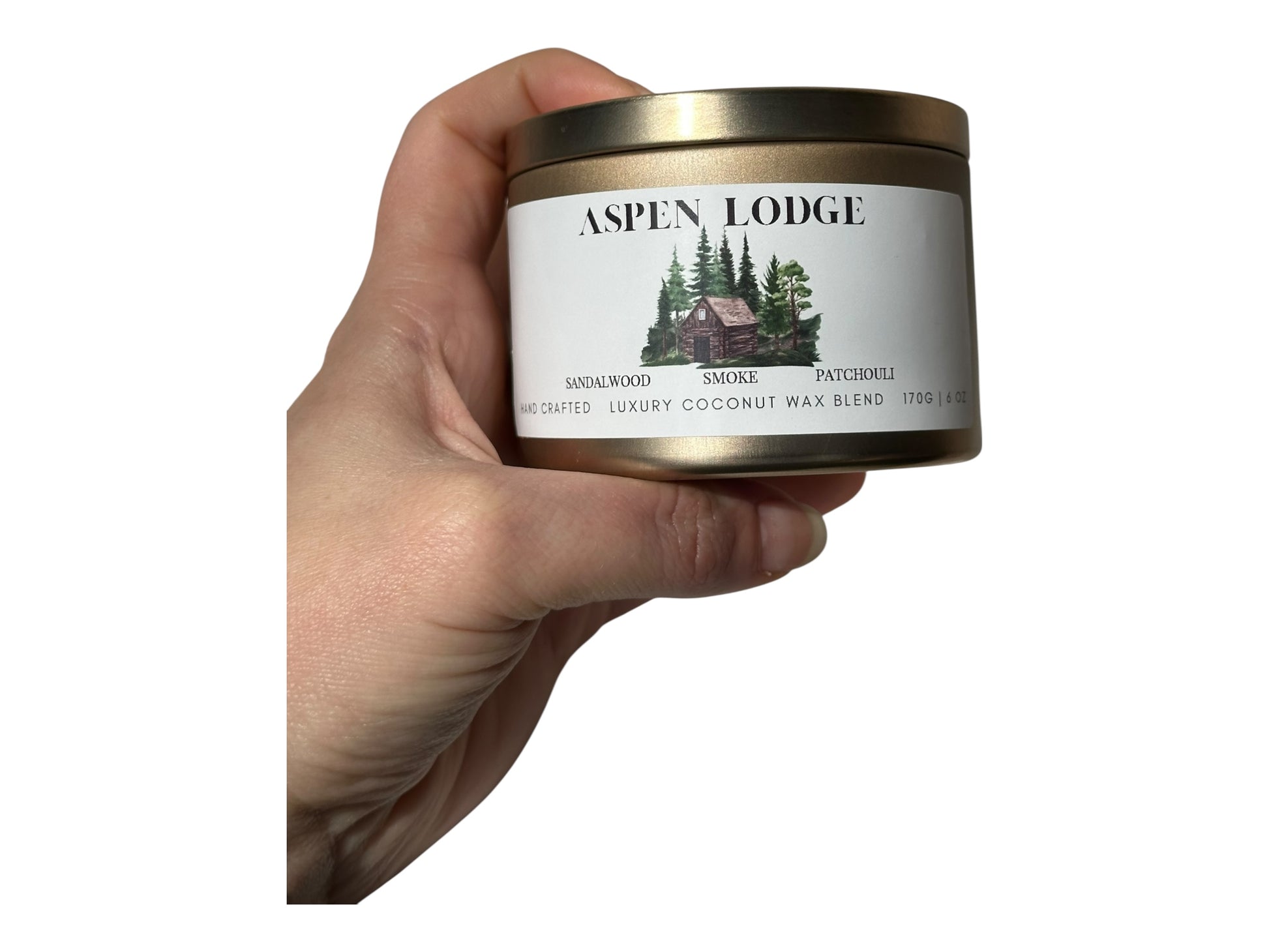 A hand holding a gold candle tin  labeled 'Aspen Lodge' on white background.