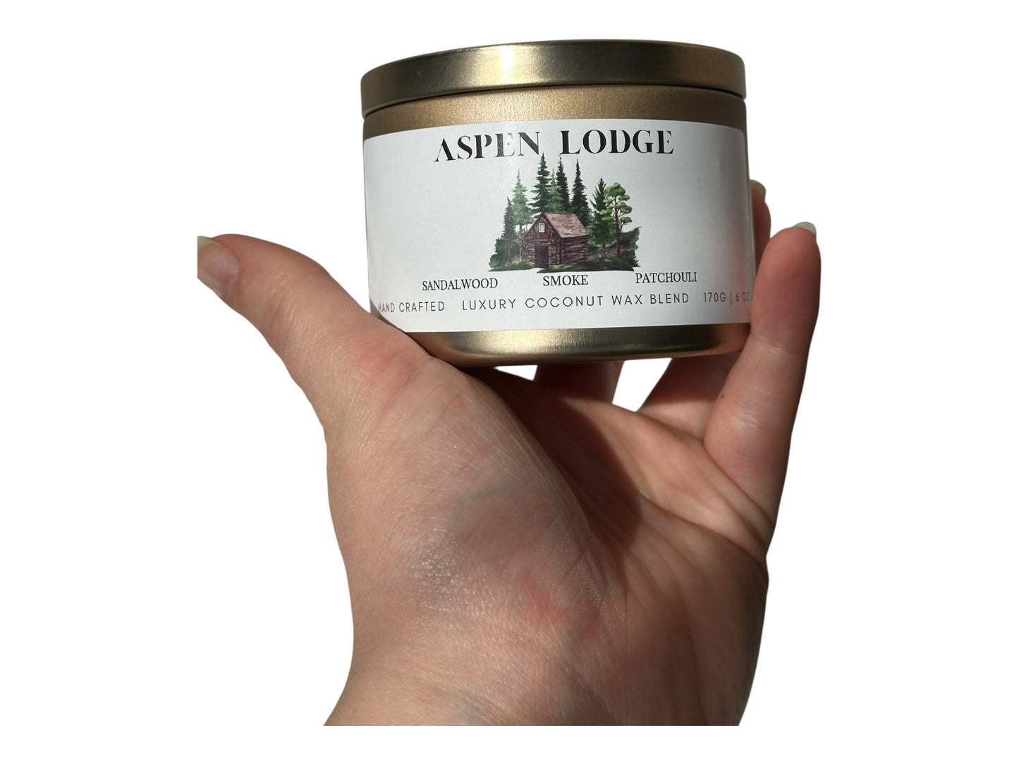 A hand holding a gold candle tin  labeled 'Aspen Lodge' against a white background.