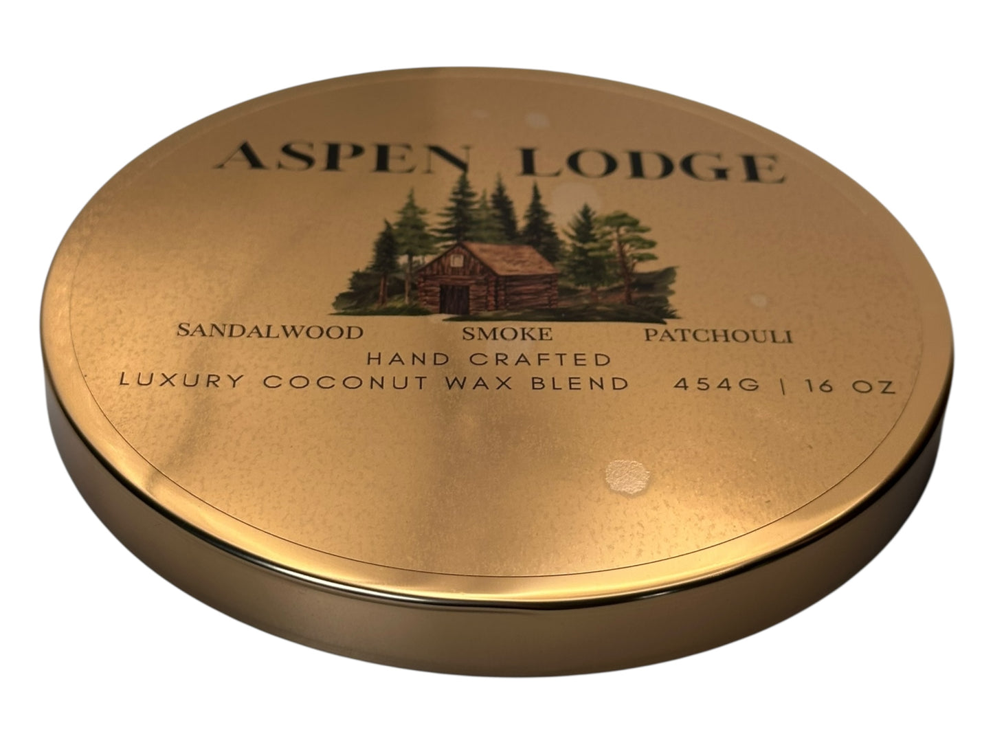 Gold candle lid labeled 'Aspen Lodge' with a cabin and tree design on a white background