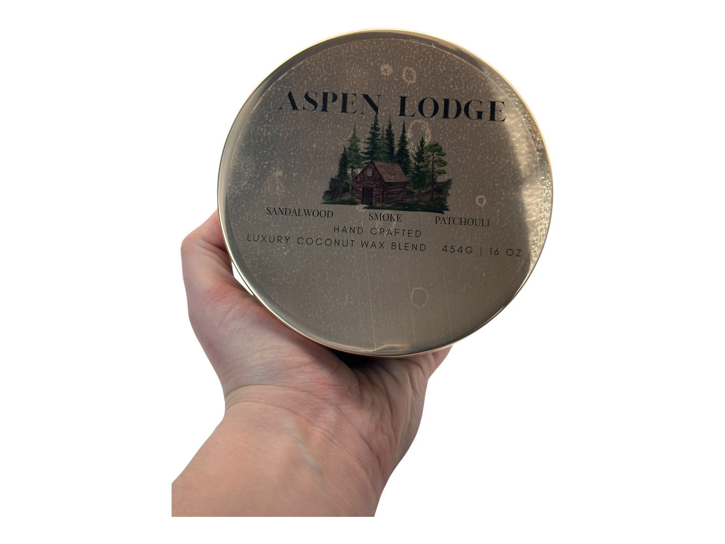 A hand holding a bronze and gold candle tin labeled "Aspen Lodge" on a white background.