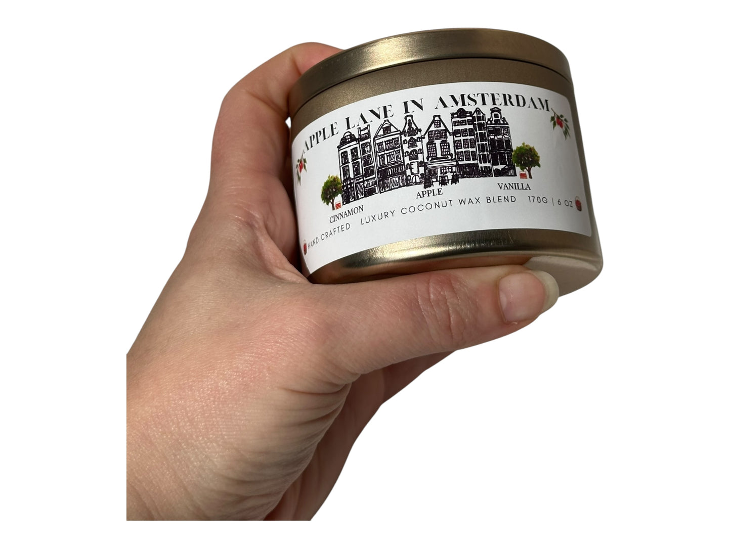 A hand holding a gold candle tin labeled 'Apple Lane in Amsterdam' against a white background.