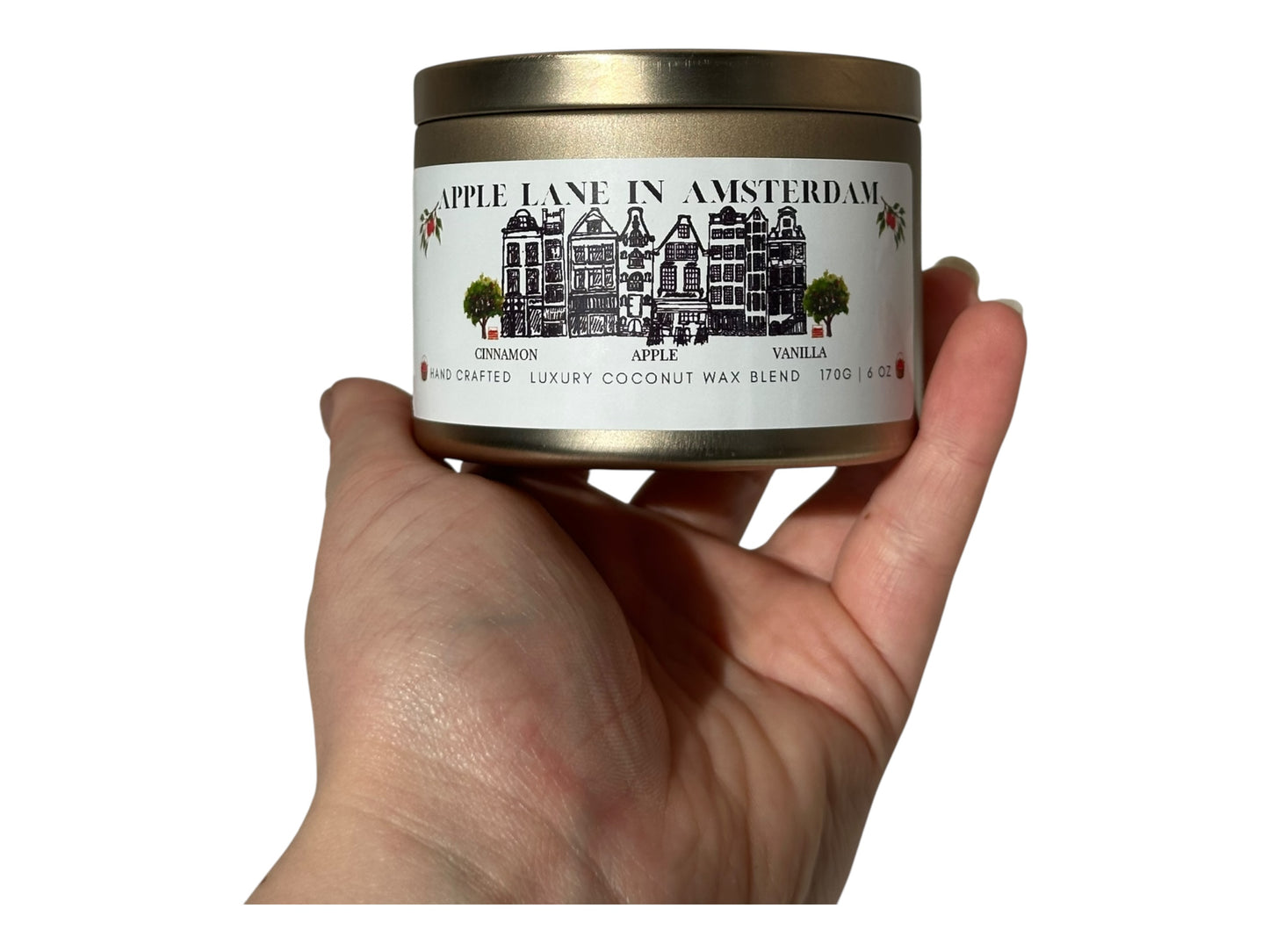 A hand holding a gold candle tin labeled 'Apple Lane in Amsterdam' against a white background.