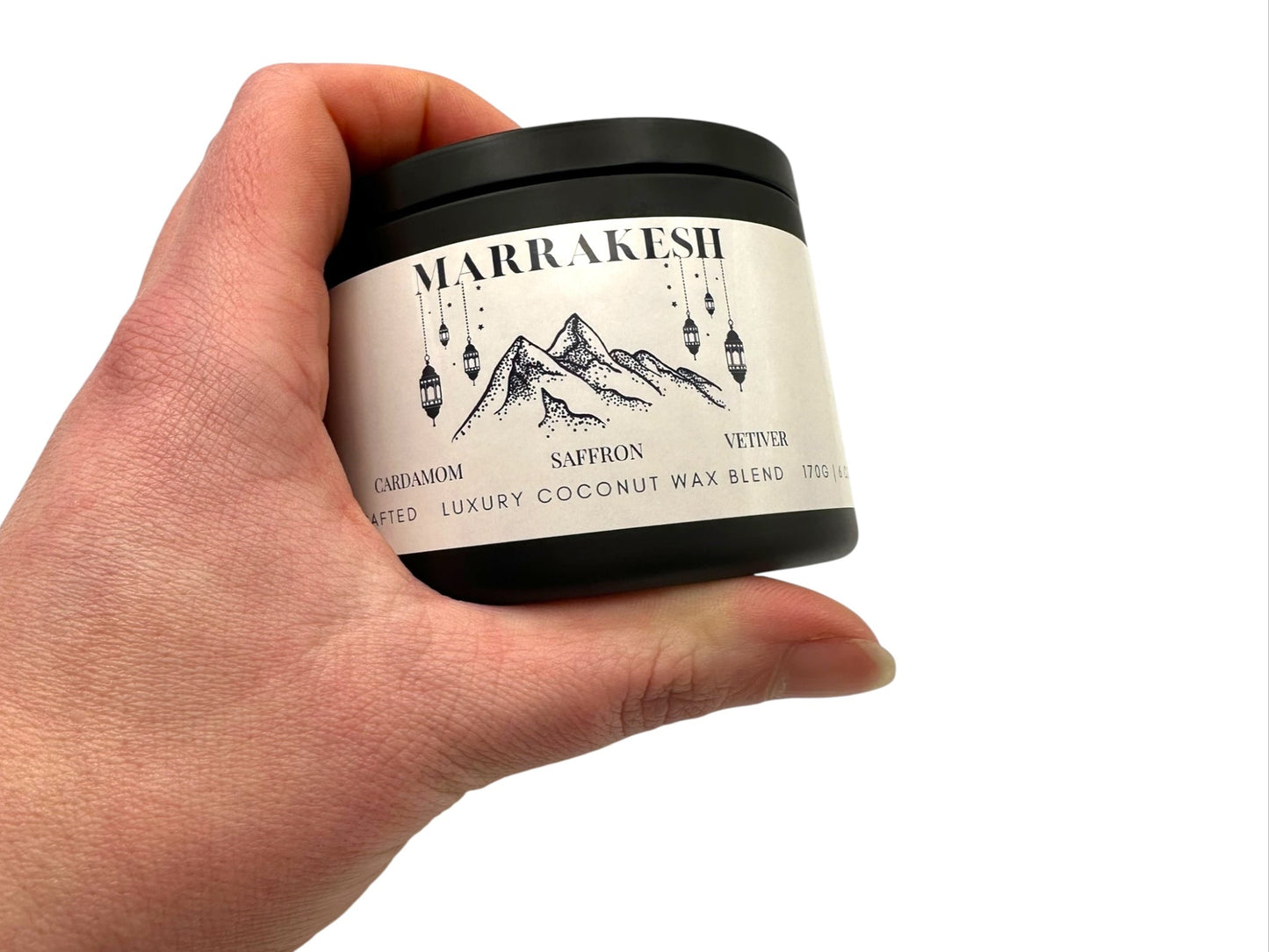 A hand holding black candle tin labeled "Marrakesh" on a white background.