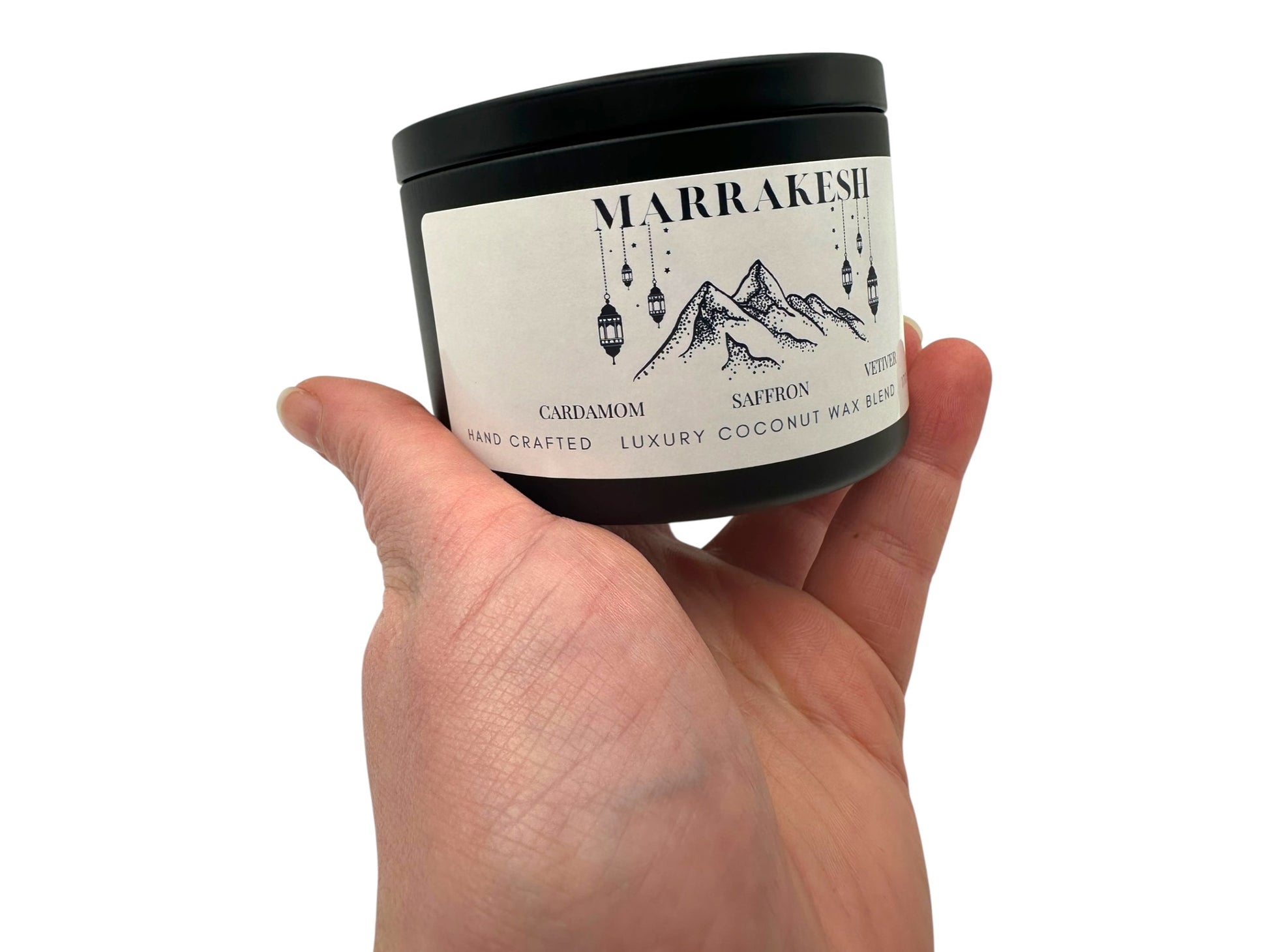 A hand holding a black candle tin labeled "Marrakesh" on a white background.