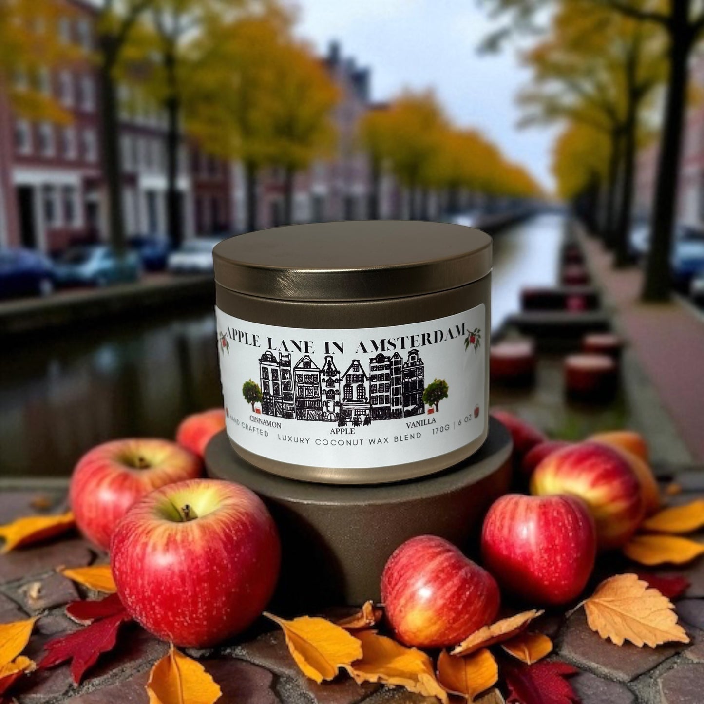 A gold candle tin labeled 'Apple Lane in Amsterdam' with apples and autumn leaves on a autumn Amsterdam background.