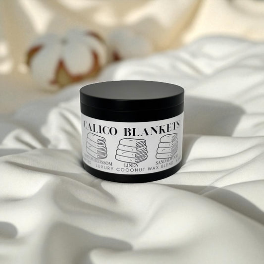 Black candle tin labeled Calico Blankets resting on a bed of fresh white linen and  cotton blossom in the background.