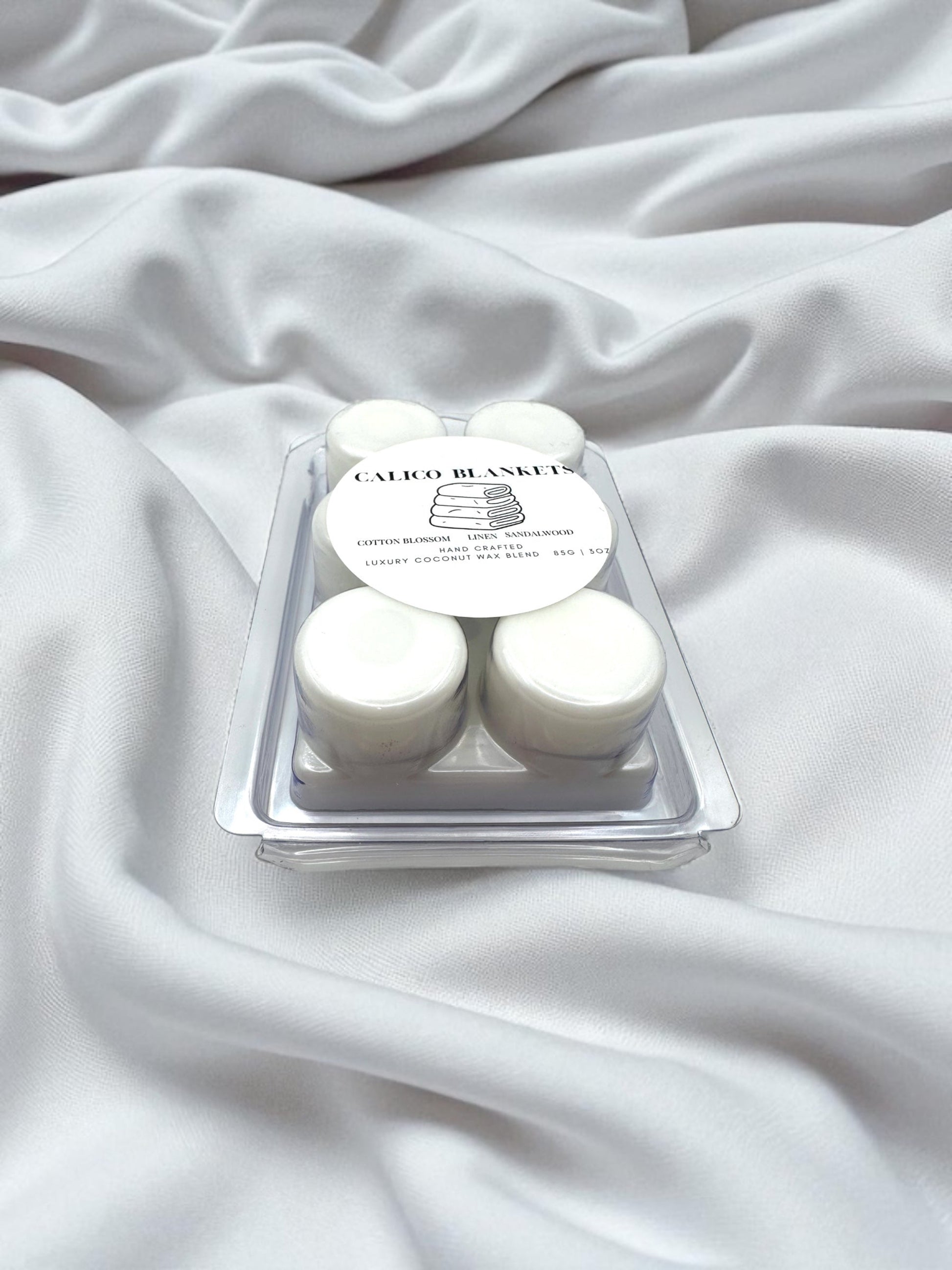 A clear wax melt clamshell labeled Calico Blankets on a bed of fresh white linen.