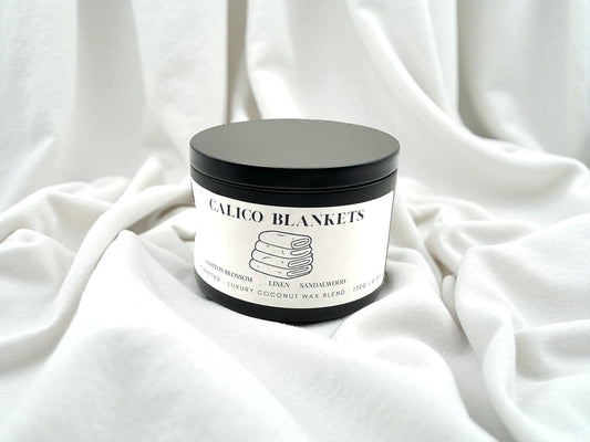 Black candle tin labeled Calico Blankets resting on a bed of white fresh linen.