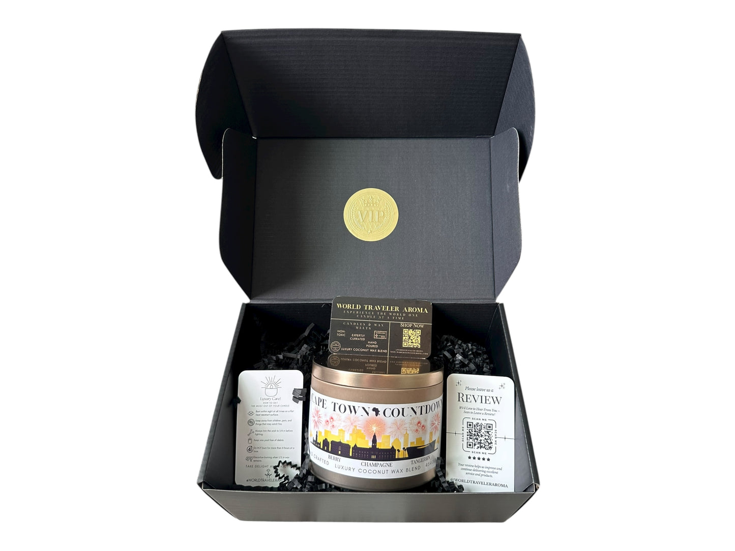 A black gift box with a gold VIP sticker on the lid inside the box a gold candle tin labeled "Cape Town Countdown" with a candle care card instructions, a please review us card, and World Traveler Aroma business card on a white background.