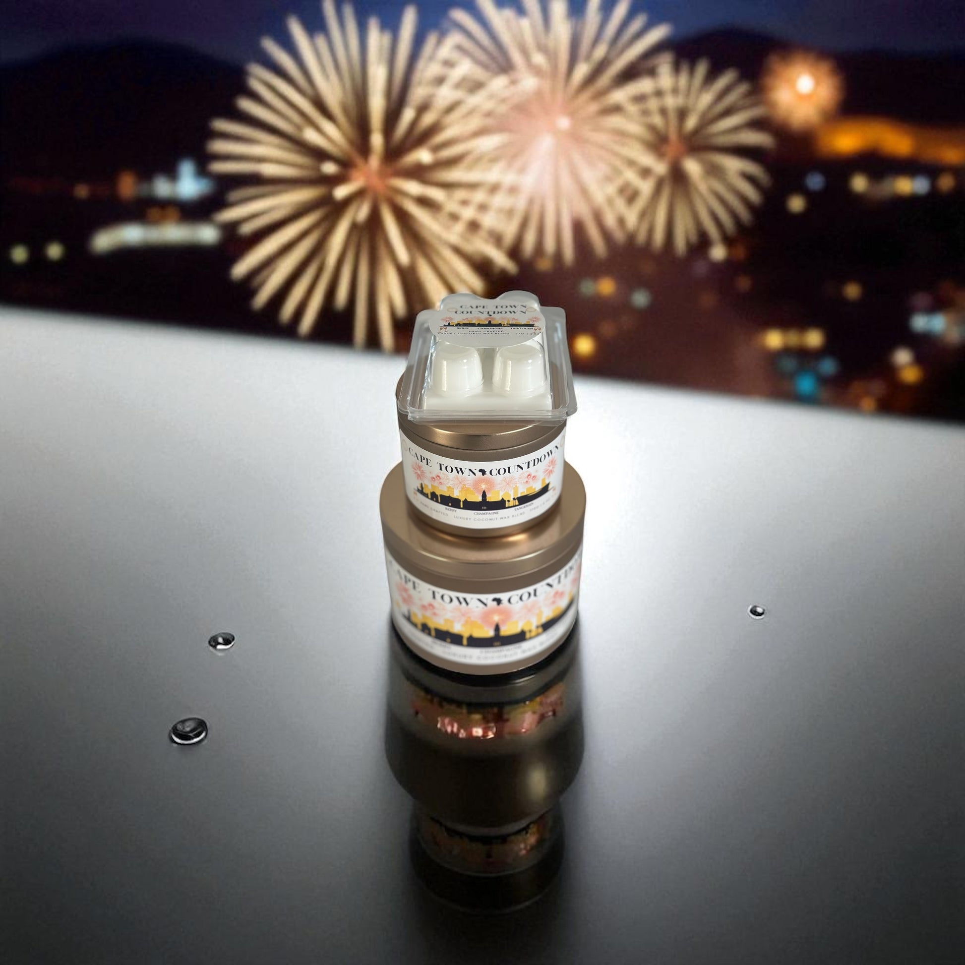 A holiday collection of gold candle tins and white wax melt labeled "Cape Town Countdown" resting on a reflective surface with Cape Town night City Scape and fireworks in the background.