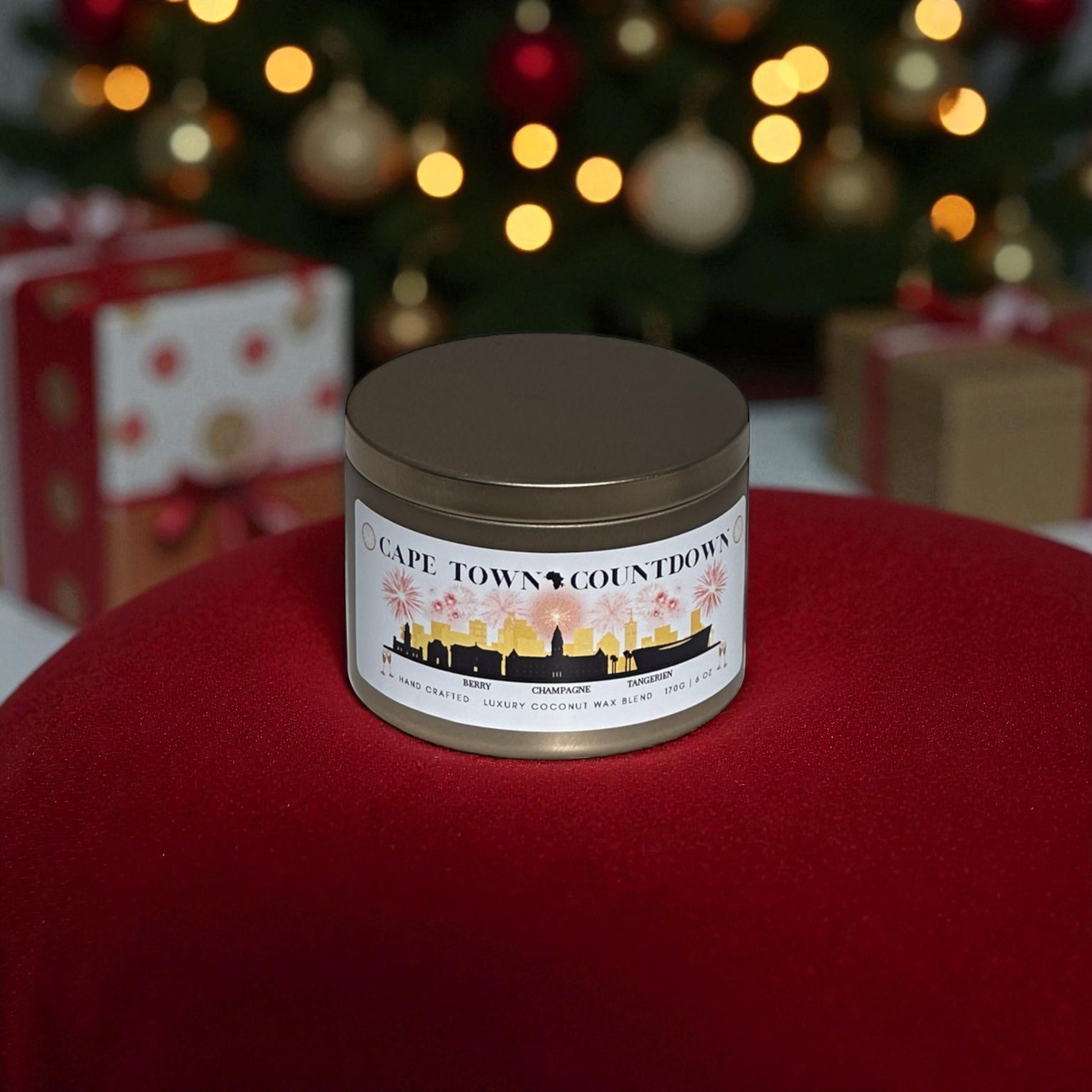 A new years gold candle tin called, "Cape Town Countdown" on a red rug with Christmas presents and a Christmas tree in the background.