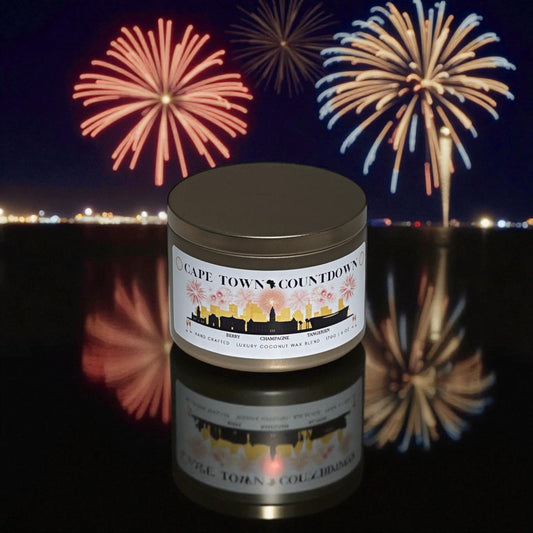 A new year's gold candle tin called, "Cape Town Countdown" on a black reflective counter top with fireworks and cityscape in the background.