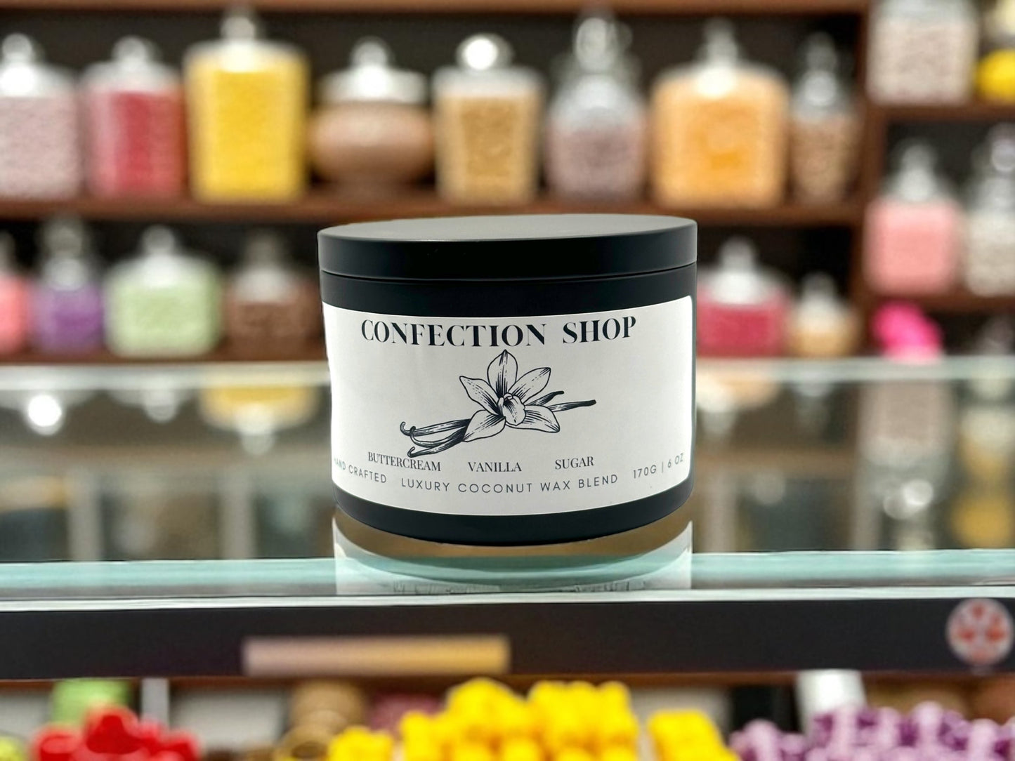 Confection Shop 6oz black candle tin in a glass counter top with candy and sweets in the background.