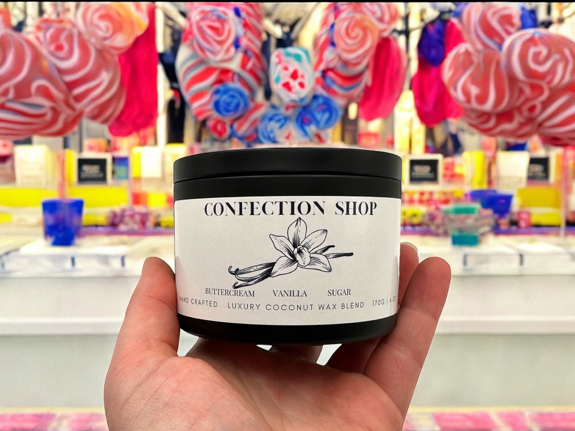 A hand holding a black candle tin labeled confection shop surrounded by candy and sweets.