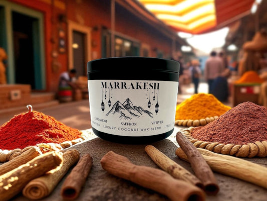 A black candle tin labeled Marrakesh resting on a concrete ledge surrounded by piles of spice with a spice market in the background.