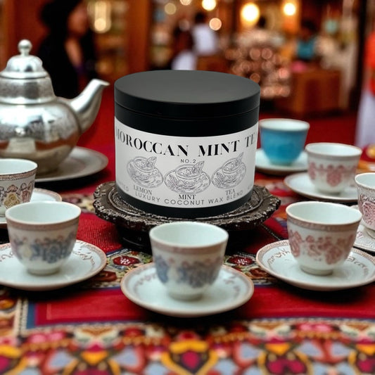 A black candle tin labeled Moroccan Mint Tea  surrounded by tea cups and a tea pot on a colorful table cloth.