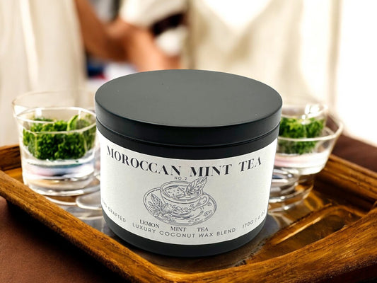 A black candle tin labeled Moroccan Mint Tea  resting on a wooden tray with two glasses of mint tea.