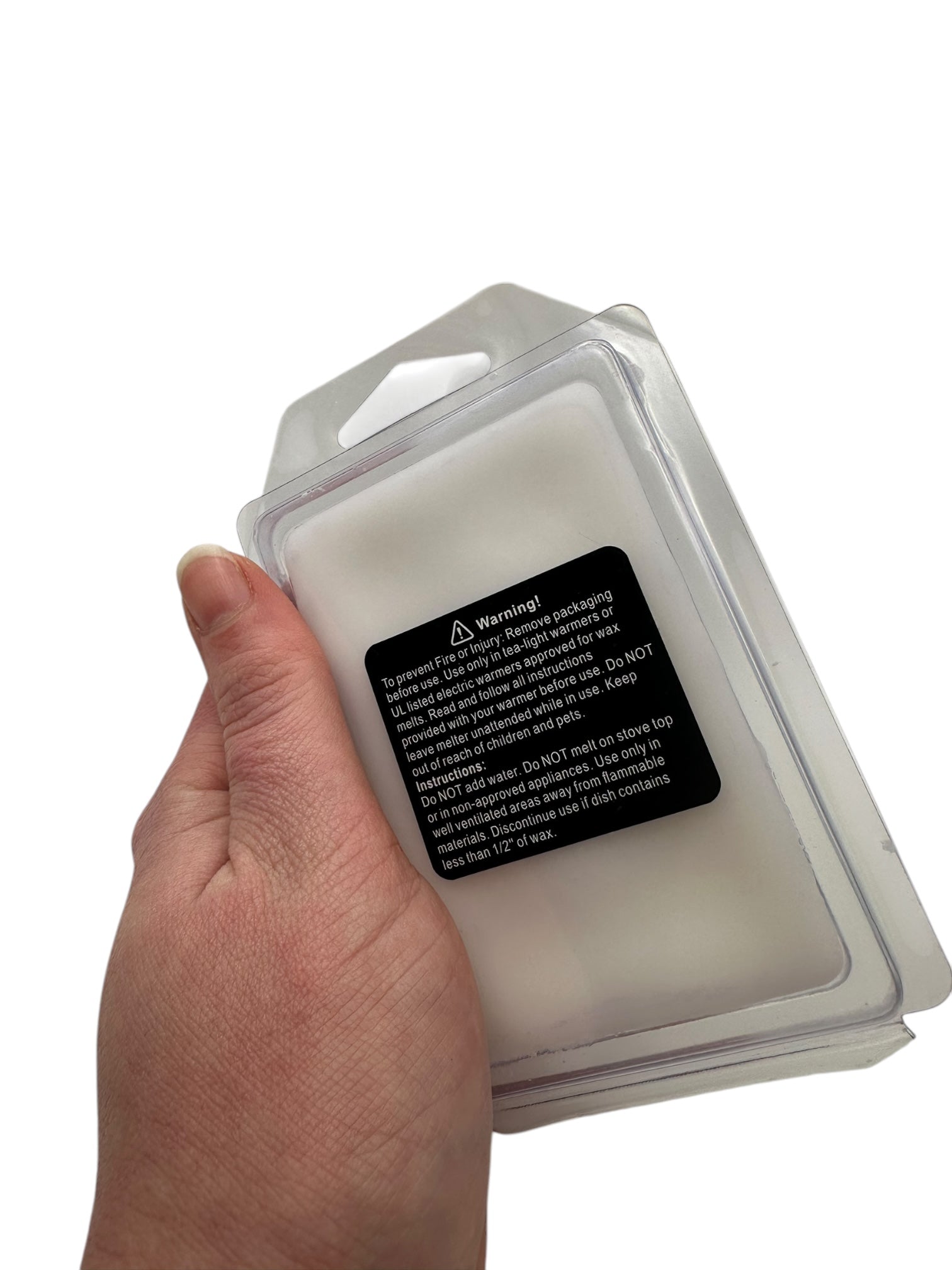 A hand holding a plastic clamshell and white wax melt with a safety warning label on the package on a white background.