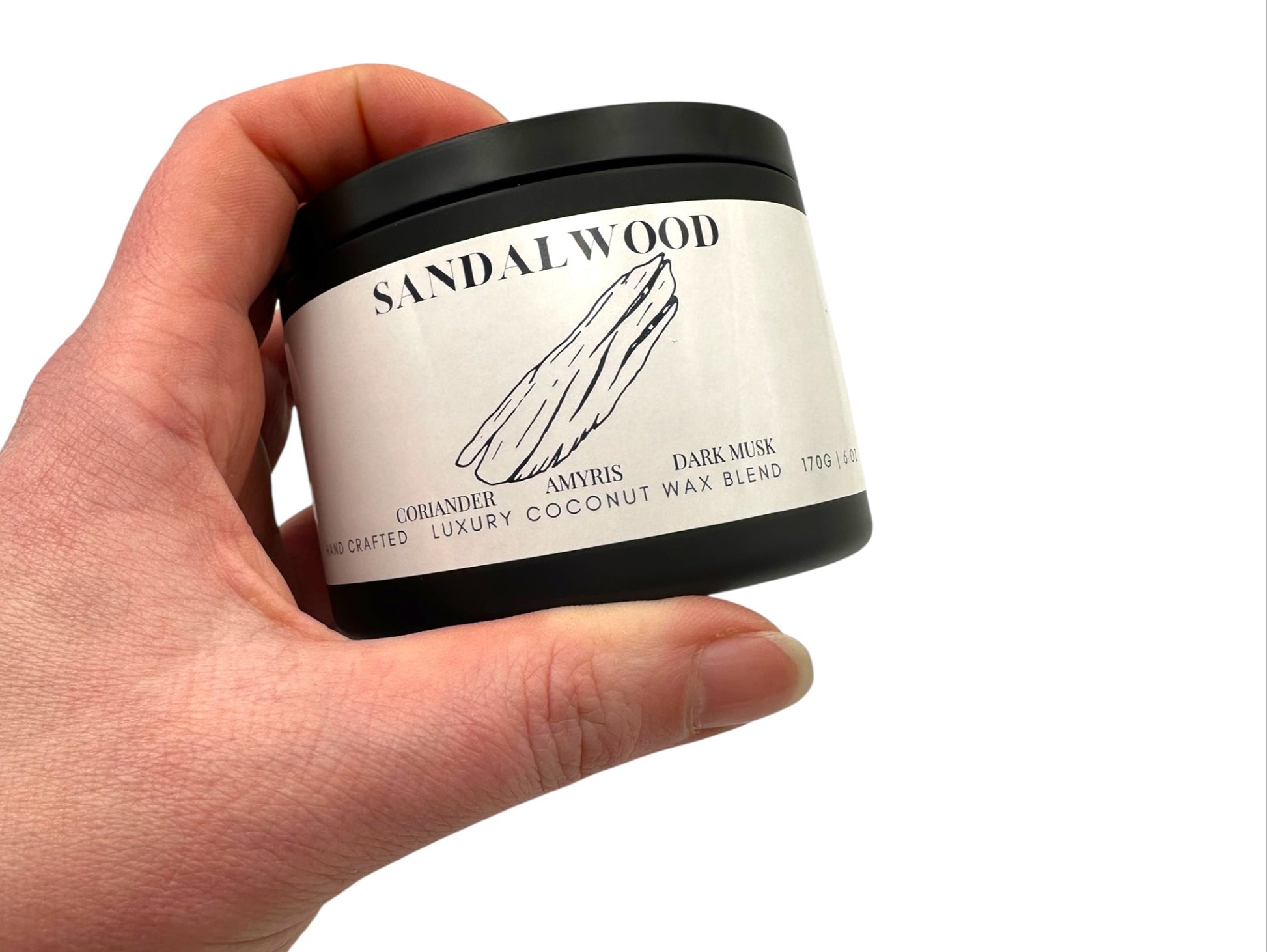 A hand holding a black candle tin labeled "Sandalwood" on a white background.