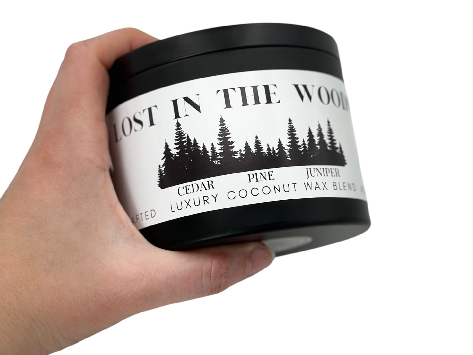 A hand holding a black candle tin labeled "Lost in the Woods" on a white background.