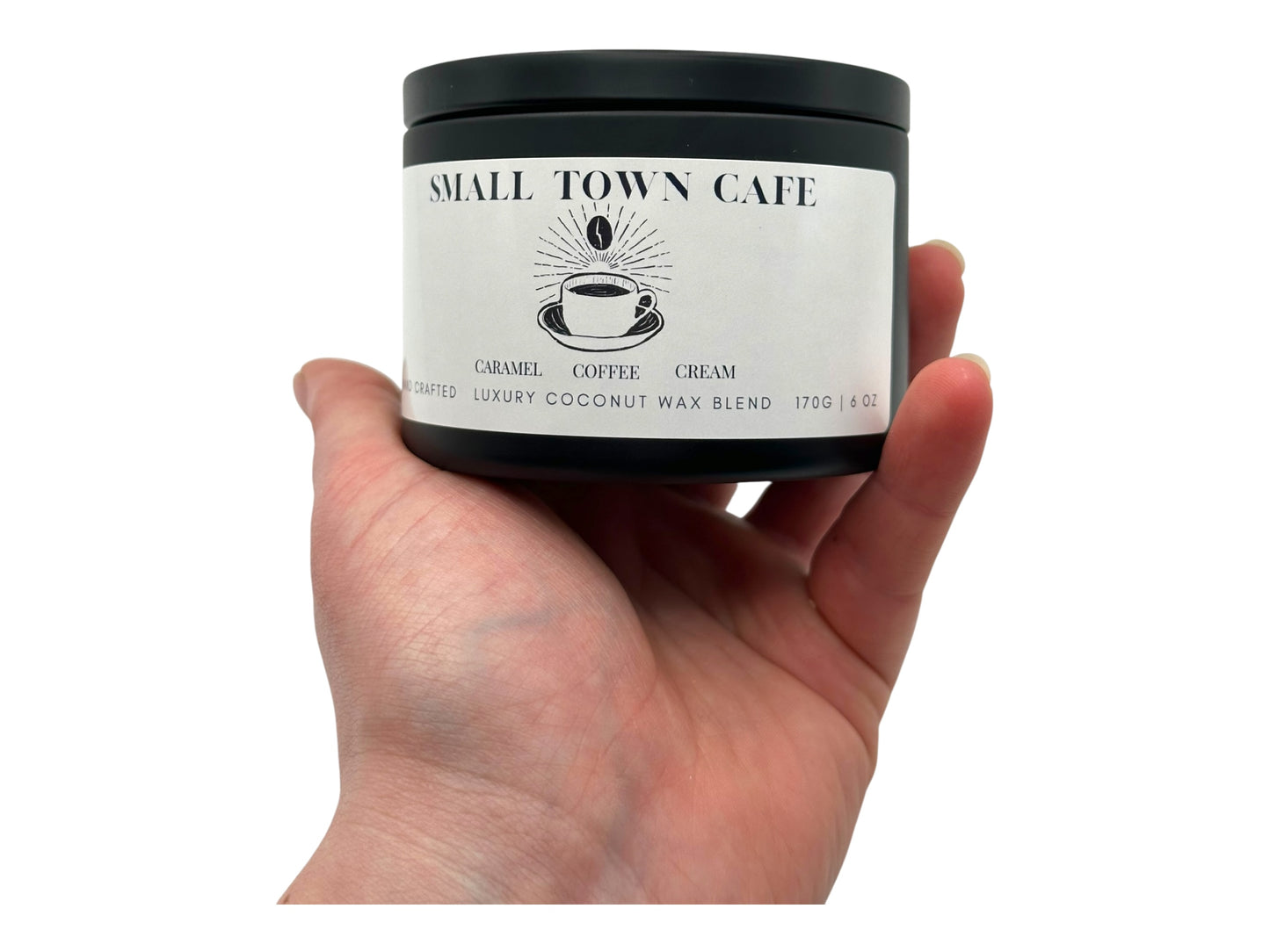 A hand holding a black candle tin labeled "Small Town Cafe" on a white background.