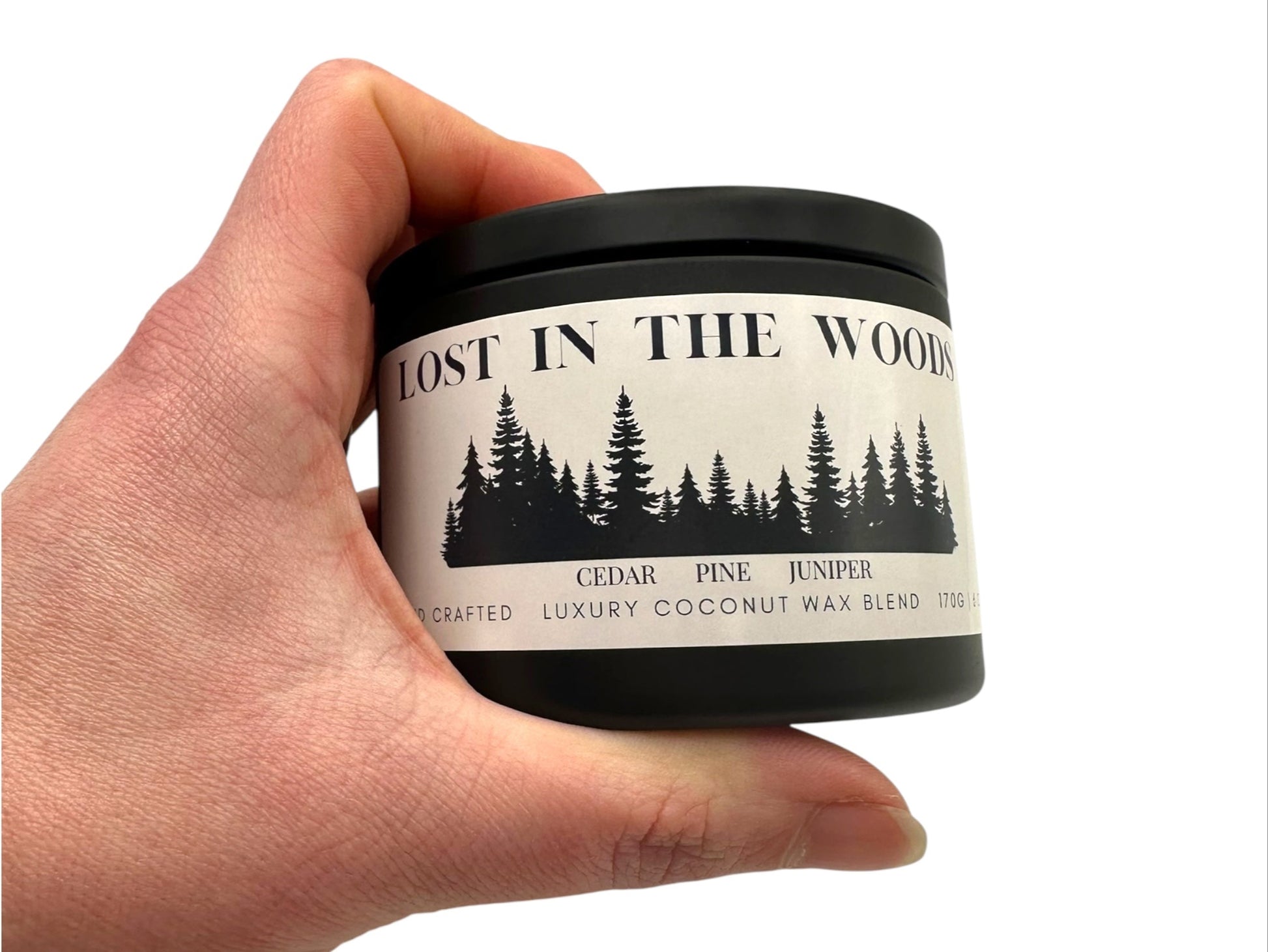 A hand holding a black candle tin labeled "Lost in the Woods" on a white background. 