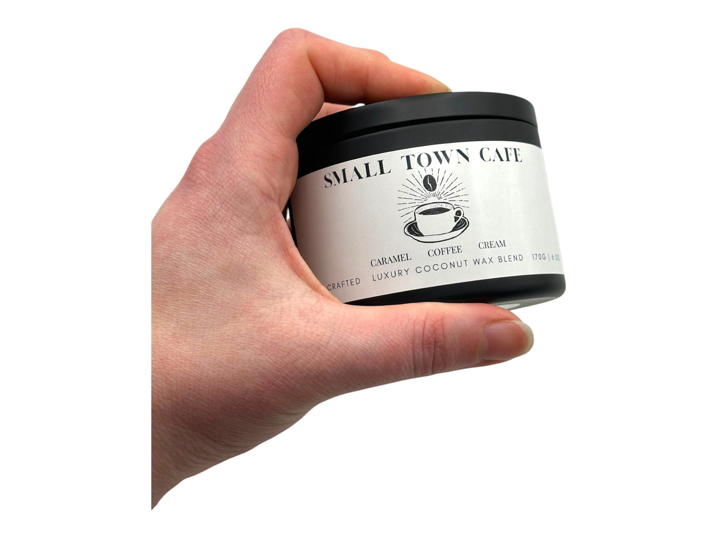 A hand holding a  black candle tin labeled "Small Town Cafe" on a white background.