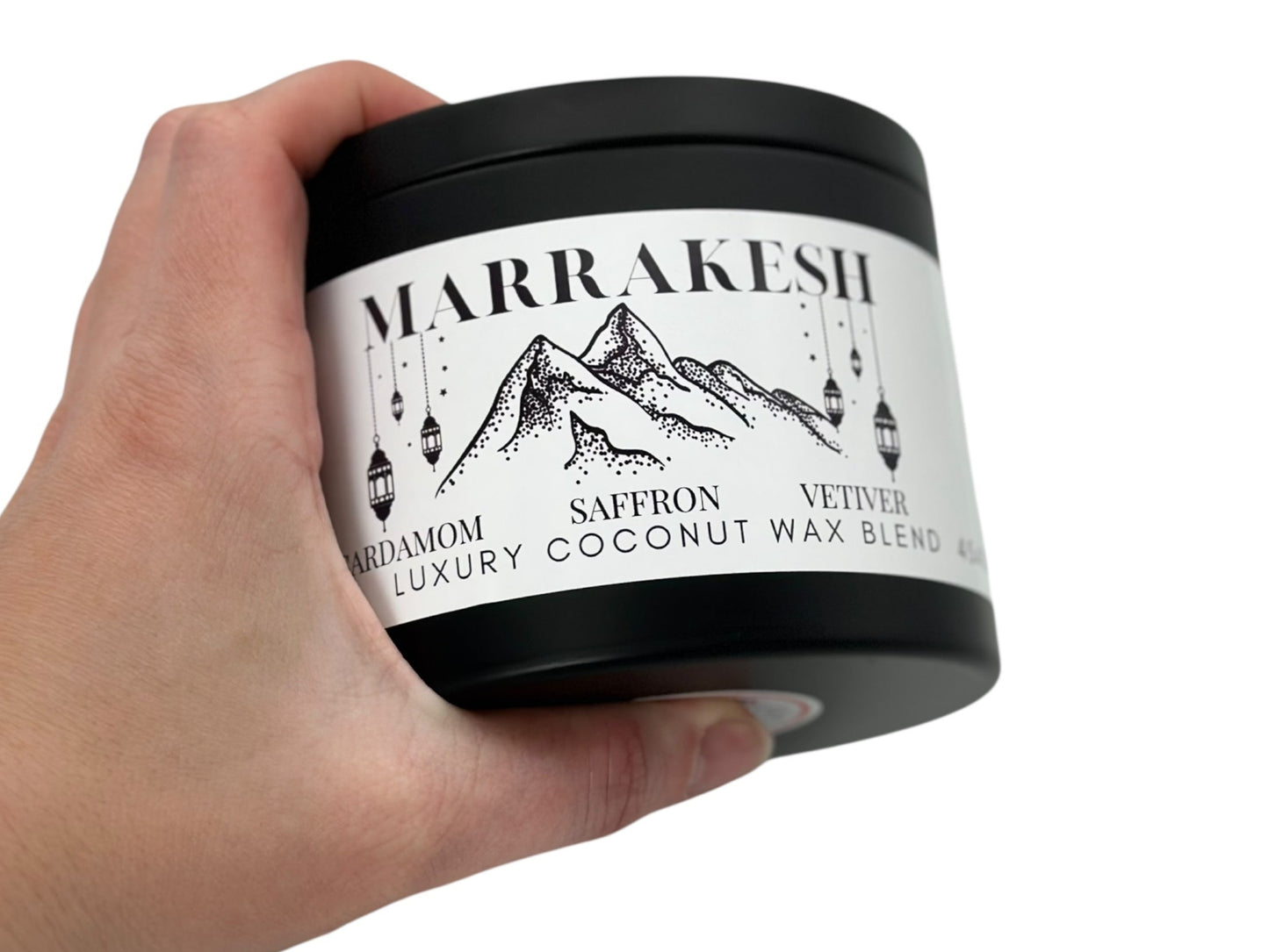 A hand holding a black candle tin labeled "Marrakesh" on a white background.