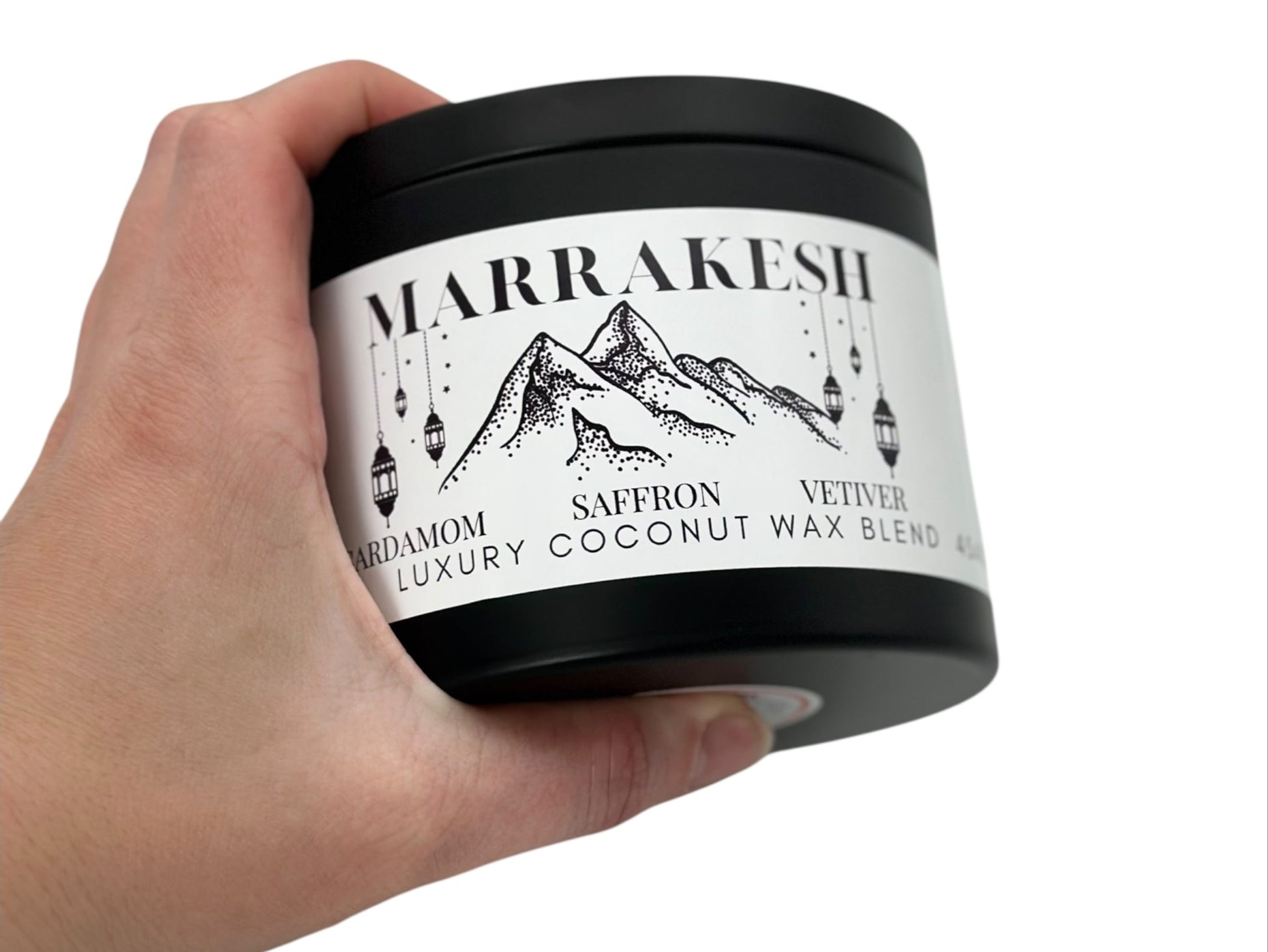 A hand holding a  black candle tin labeled "Marrakesh" on a white background.