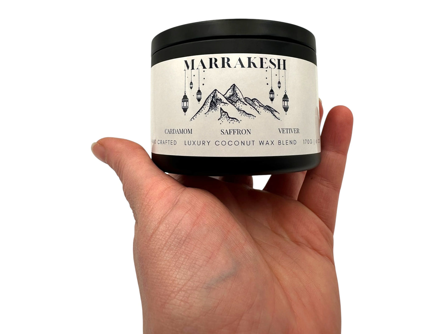 A hand holding a black candle tin labeled "Marrakesh" on a white background. 