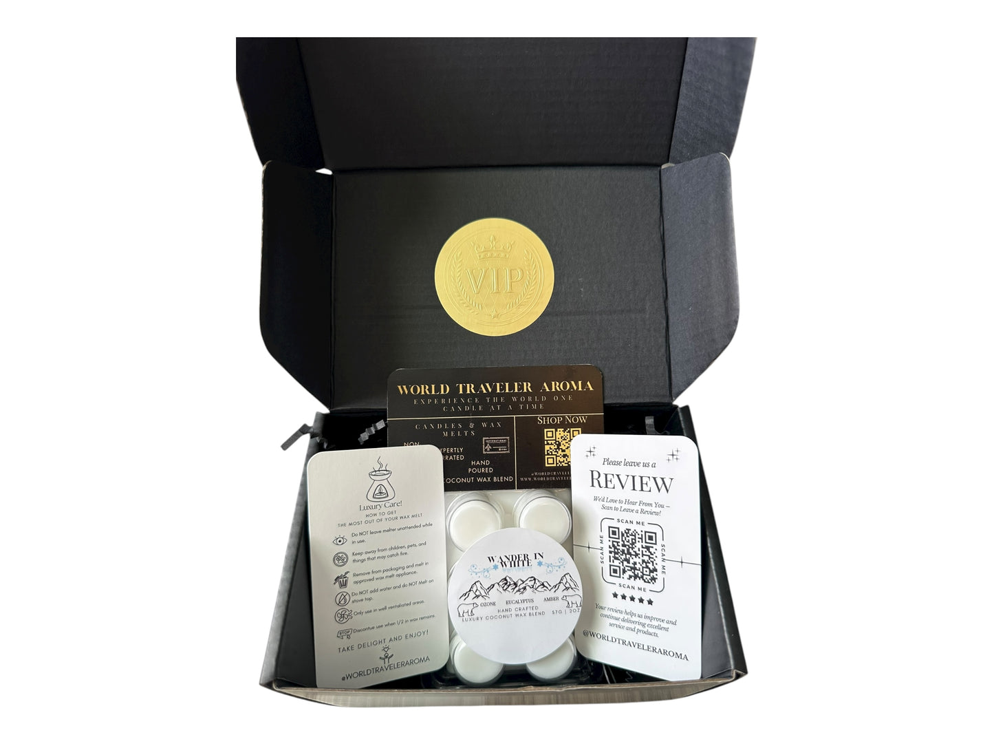 A black gift box with a gold VIP sticker on the lid with holiday wax melt labeled "Wander in White" a how to care card, please review card, and World Traveler Aroma Business Card on a white background.