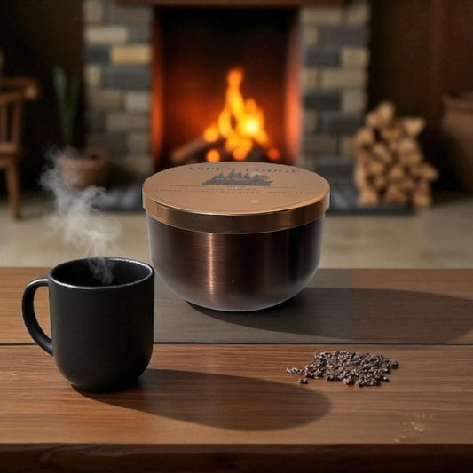 A bronze and gold candle tin and black mug on a wooden table in front of a fireplace.