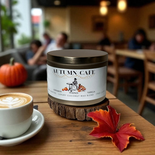 A candle labeled 'Autumn Cafe' on a wooden surface with a cup of coffee and a pumpkin in the background.