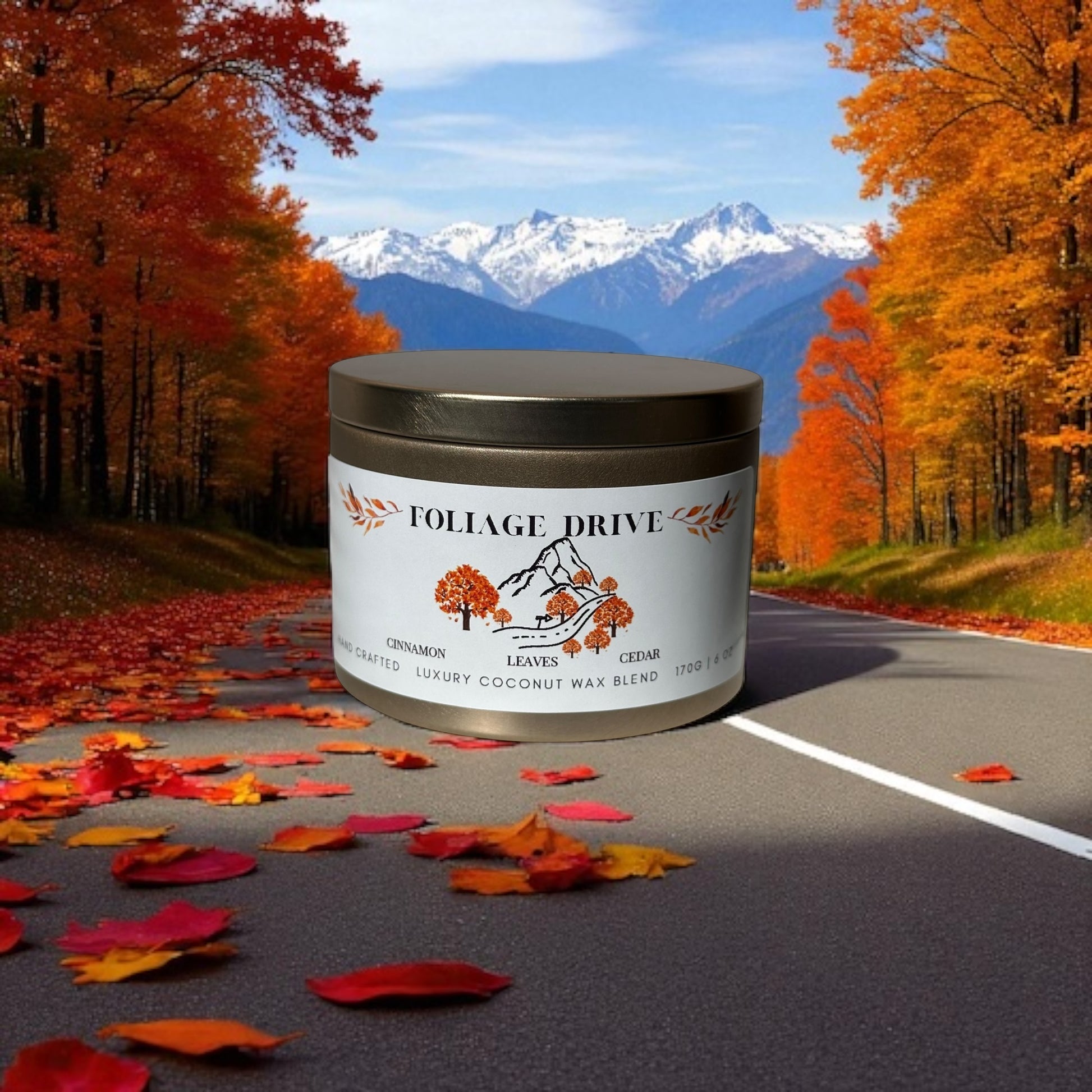 A gold candle tin labeled 'Foliage Drive' on a road with autumn leaves and mountains in the background.