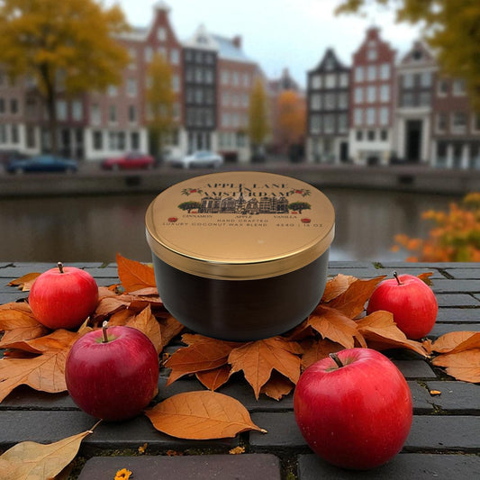 A bronze and gold candle tin on a brick road  with red apples and autumn leaves, blurred canal and buildings in the background.