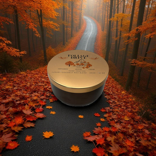 A bronze and gold candle tin labeled "Foliage Drive" on a road flanked by trees with autumn foliage.