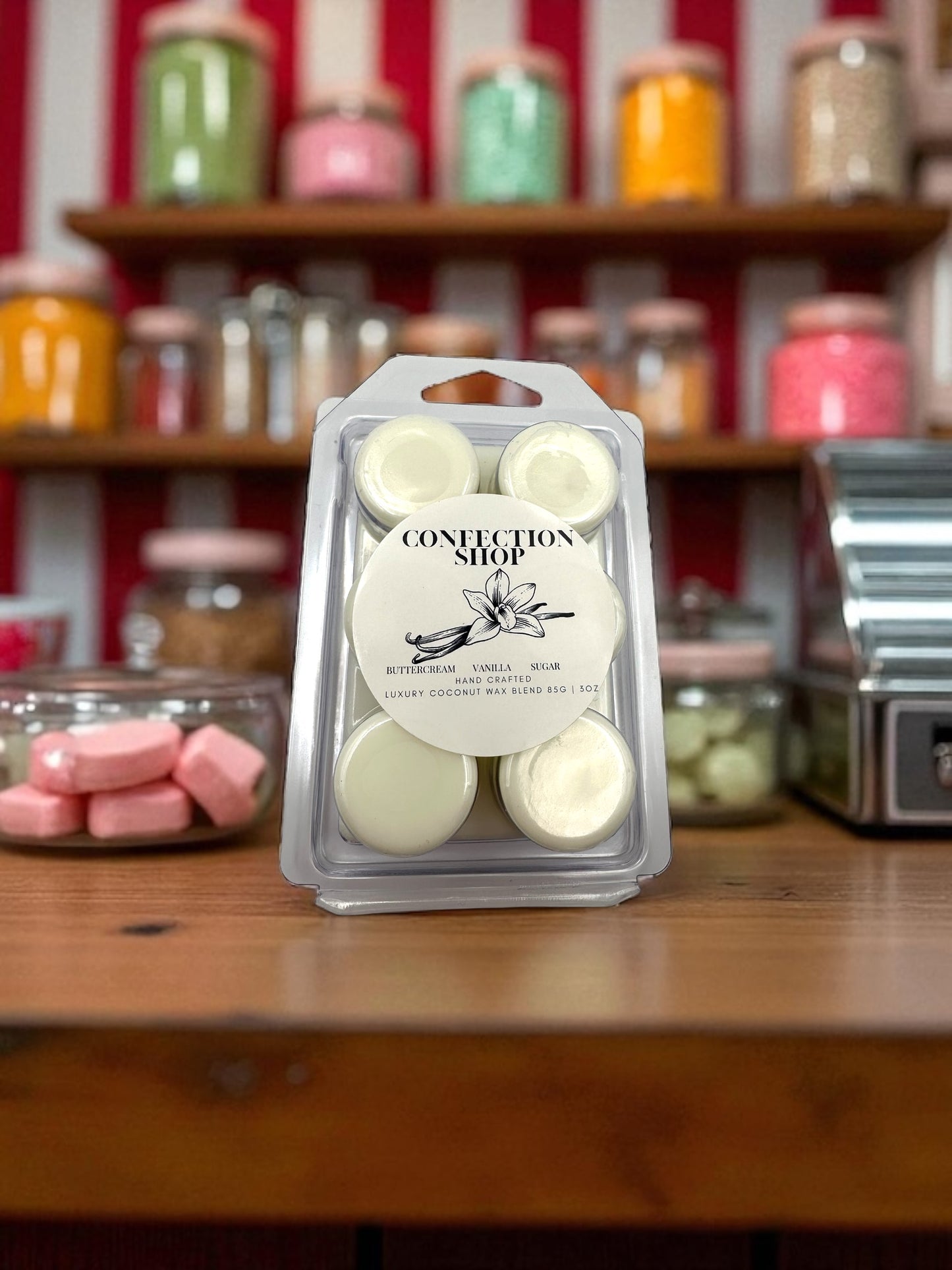A white wax melt in a clear plastic clamshell labeled “Confection Shop” sitting on a wood counter top in a candy store.