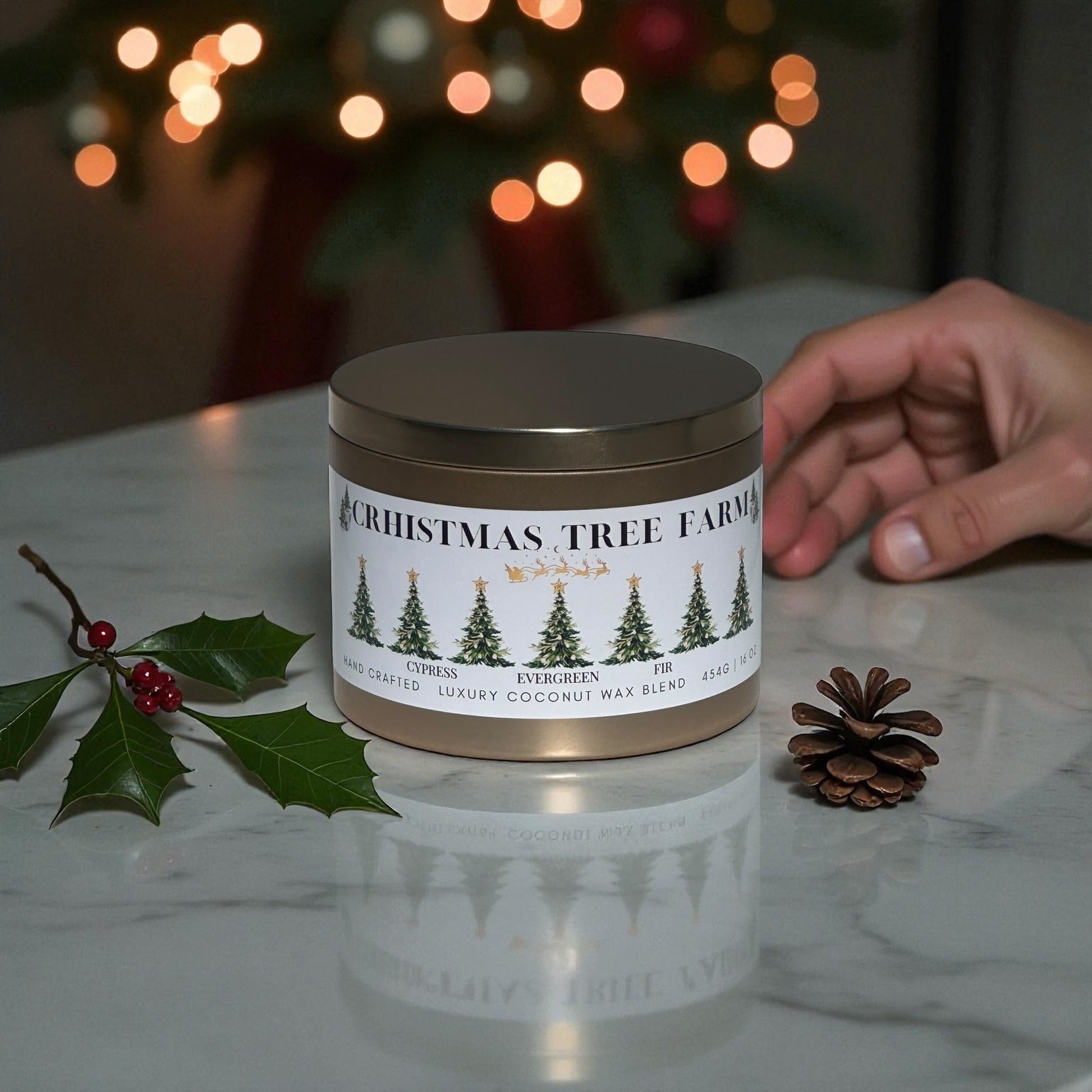 A gold candle tin labeled, "Christmas Tree Farm" sitting on a white marble counter top with a stem of holy a mini pinecone and a had sitting next to the candle with a Christmas tree in the background.