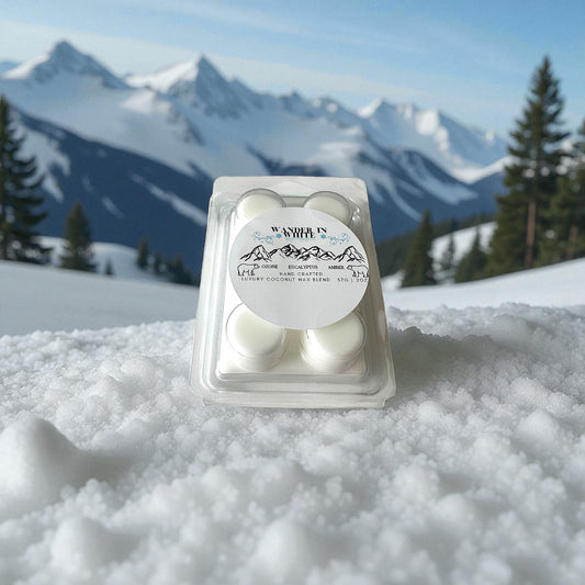 A holiday wax melt labeled "Wander in White" sitting on fresh snow with a snow covered mountains and pines in the background.