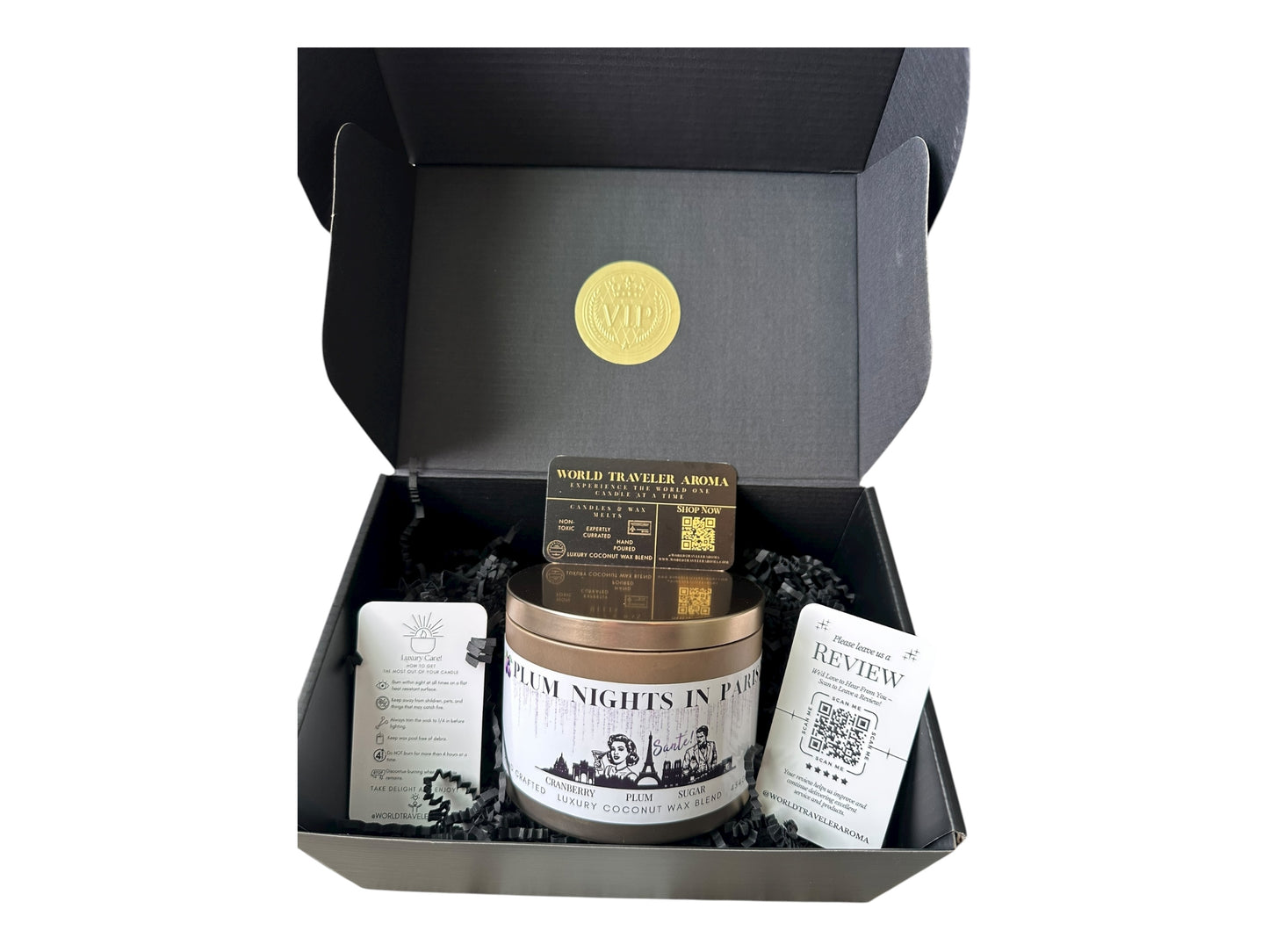A black gift box with a gold VIP sticker on the lid inside the box a gold candle tin labeled "Plum Nights in Paris" a candle care care instructions, a please review us card, and a World Traveler Aroma business card on a white background.