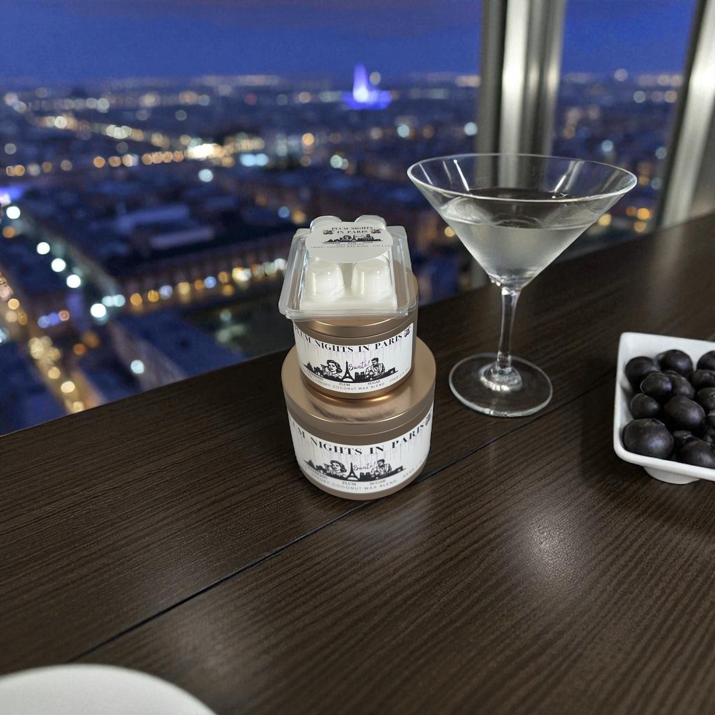 A Christmas collection of gold candle tins and white wax melts labeled "Plum Nights in Paris" resting on a wooden surface with a Martini and plater of plums next to the collection with a night time city scape in the background.